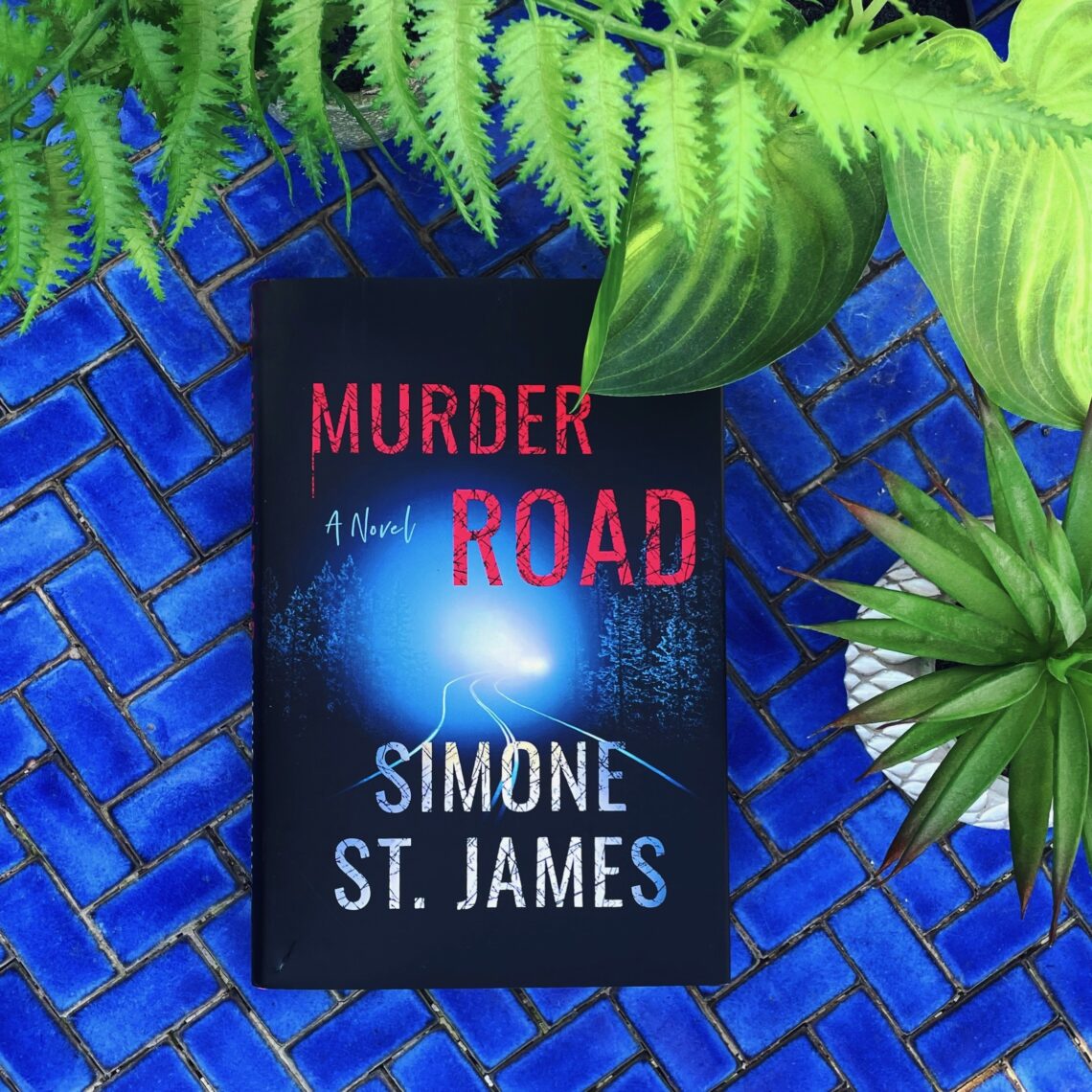 Murder Road is a supernatural mystery by Simone St James showing a dark road with a car driving towards the viewer with bright headlights